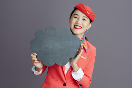 smiling elegant asian female flight attendant in red skirt, jacket and hat uniform showing blank...