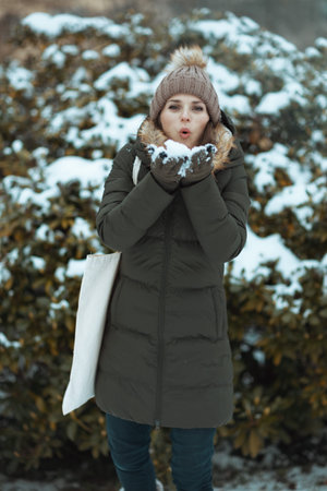 modern woman in green coat and brown hat outdoors in the city park in winter with mittens and beanie...