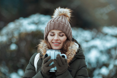 smiling modern 40 years old woman in green coat and brown hat outdoors in the city park in winter with mittens, cup of coffee and beanie hat.の写真素材