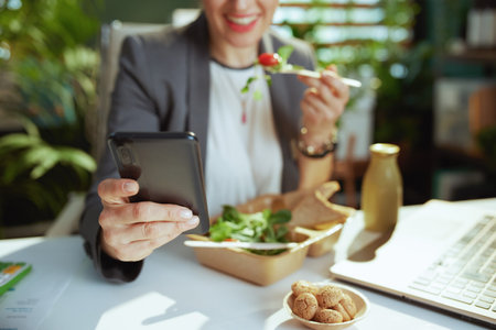 Sustainable workplace. happy modern middle aged small business owner woman in a gray business suit in modern green office with laptop eating salad and using smartphone app.の写真素材