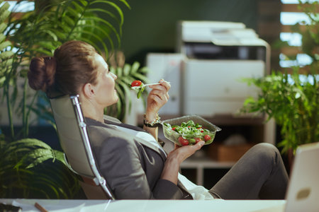 Sustainable workplace. relaxed modern business woman in a gray business suit in modern green office with laptop eating salad.の写真素材