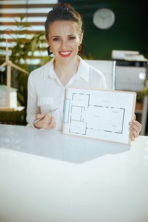 Time to move on. smiling elegant 40 years old woman real estate agent in modern green office in white blouse with clipboard and building plan.の写真素材