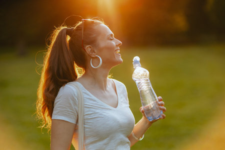 Summer time. smiling elegant middle aged woman in white shirt with bottle of water in the meadow in...