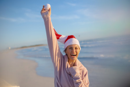 smiling modern woman in cozy sweater with striped Christmas hat rejoicing at the beach in the evening.の写真素材