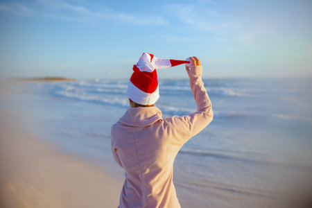 Seen from behind modern middle aged woman in cozy sweater with striped christmas hat having fun time at the beach in the evening.の写真素材