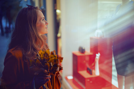Hello September. elegant middle aged woman in orange trench coat with autumn yellow leaves near shop...