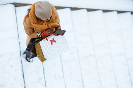 Upper view of woman in brown hat and scarf in camel coat with shopping bags using smartphone app outside in the city in winter.の写真素材