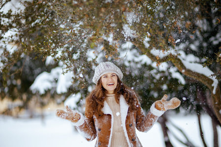 smiling stylish woman in brown hat and scarf in sheepskin coat with mittens catching snow outside in...