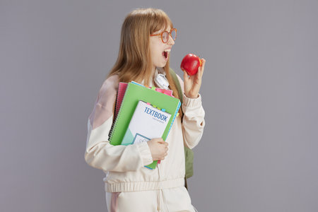 happy modern young woman in beige tracksuit with apple, workbooks, headphones, textbook and eyeglasses against grey.の写真素材