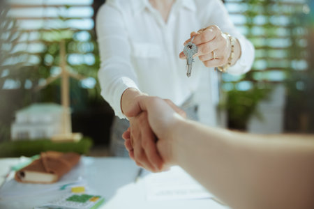 Sustainable real estate business. Closeup on modern 40 years old woman realtor in modern green office in white blouse with keys and client shaking hands.の写真素材