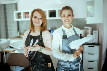 Portrait of happy two 40 year old women hairdressers in aprons in modern beauty salon with hair straightener and hair dryer.の写真素材