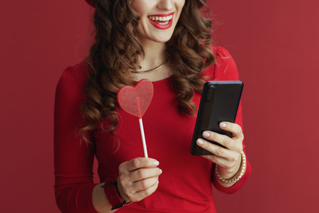 Happy Valentine. Closeup on woman isolated on red background with heart shaped candy on stick using smartphone app.の写真素材