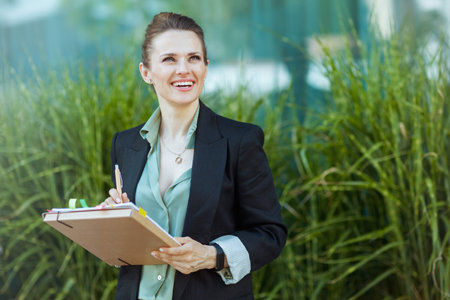 happy modern middle aged business woman near office building in black jacket with documents and folder.の写真素材