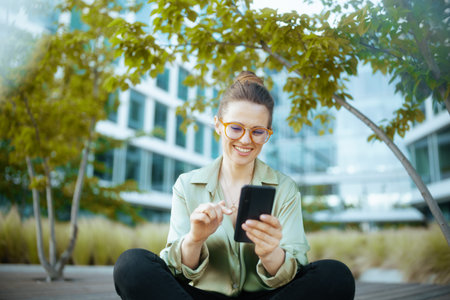 smiling modern business woman in business district in green blouse and eyeglasses with smartphone.の写真素材