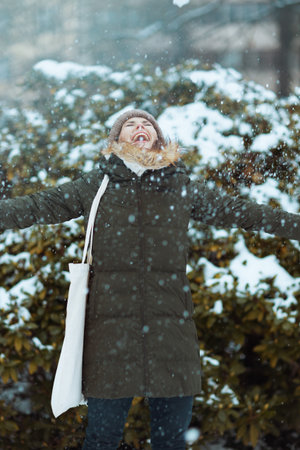 smiling modern woman in green coat and brown hat outdoors in the city park in winter with mittens...