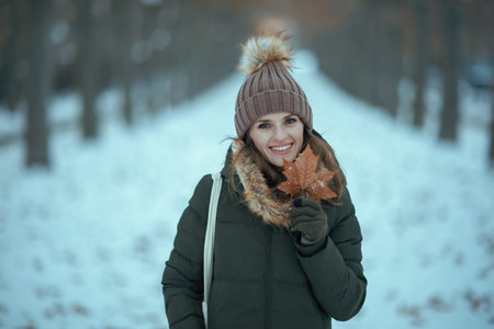 happy modern middle aged woman in green coat and brown hat outdoors in the city park in winter with...