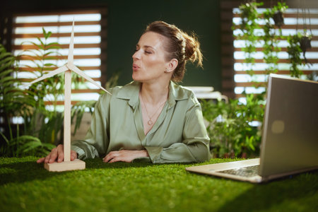 Sustainable business. smiling young business woman in green blouse in modern green office with laptop and windmillの写真素材