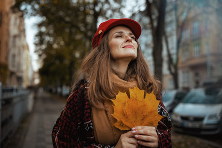 Hello autumn. relaxed elegant middle aged woman in red hat with autumn leaves and scarf in the city.の写真素材