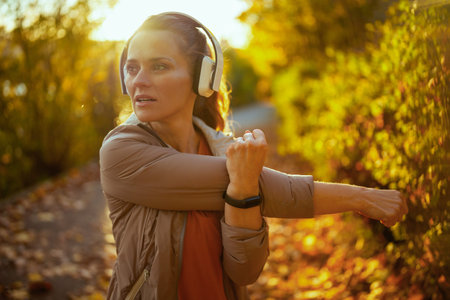 Hello autumn. stylish 40 years old woman in fitness clothes in the park with headphones stretching.
