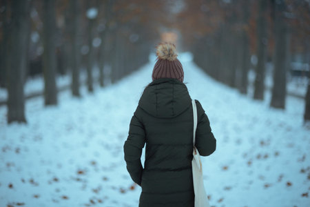 Seen from behind modern woman in green coat and brown hat outdoors in the city park in winter with beanie hat.の写真素材