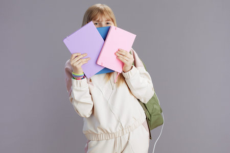 trendy teenage girl in beige tracksuit with backpack and workbooks peeking out of notebooks isolated...