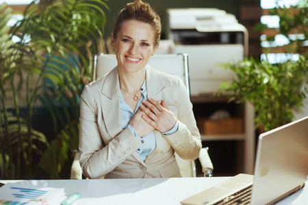 Portrait of happy modern middle aged woman employee in a light business suit in modern green office with laptop and crossed arms on chest.の写真素材