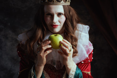 insidious medieval queen in red dress with green apple, white collar and crown on dark gray background.の写真素材