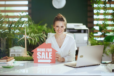Sustainable real estate business. smiling modern female real estate agent in modern green office in white blouse with home for sale sign and laptop.の写真素材