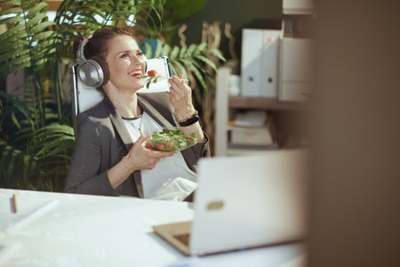 Sustainable workplace. smiling modern accountant woman in a gray business suit in modern green office with laptop eating salad and listening to the music with headphones.の写真素材