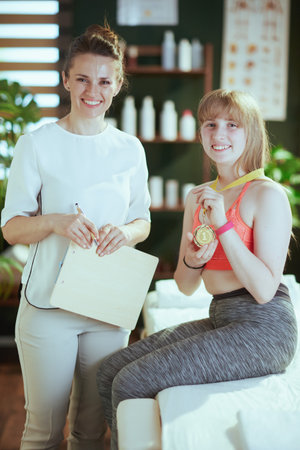 Healthcare time. happy massage therapist woman in massage cabinet with clipboard, teenage client and golden medal.の写真素材