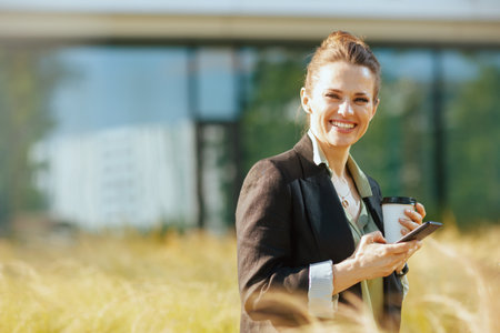 Portrait of happy modern middle aged woman employee near office building in black jacket with cup of coffee using smartphone.の写真素材