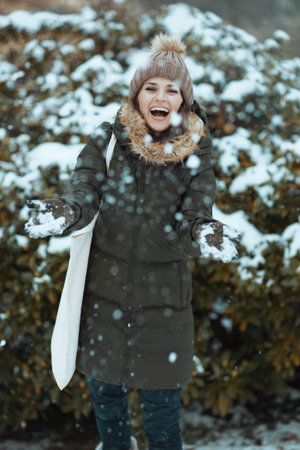 smiling modern woman in green coat and brown hat outdoors in the city park in winter with mittens and beanie hat playing with snow near snowy branches.の写真素材