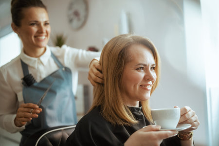 40 years old hair salon employee in modern beauty salon cutting hair with scissors and relaxed client with cup of tea.の写真素材