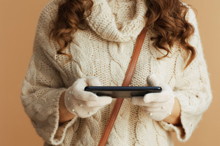 Hello winter. Closeup on woman in beige sweater, mittens and hat on beige background using smartphone applications.の写真素材