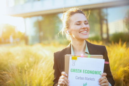 smiling modern middle aged business woman near business center in black jacket with documents, green economy plans, folder and wireless headphones.の写真素材