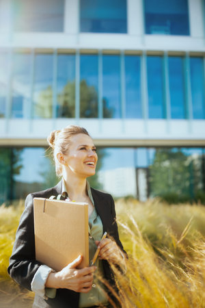 happy modern middle aged woman worker near office building in black jacket with documents and folder.の写真素材
