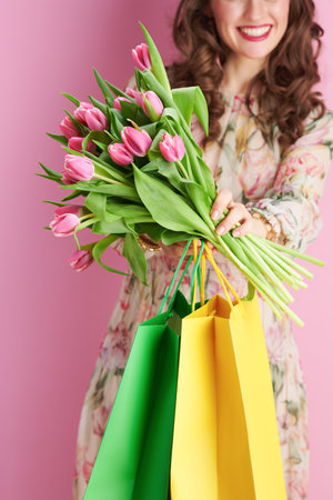 Closeup on smiling middle aged woman in floral dress with tulips bouquet and shopping bags isolated on pink background.の写真素材