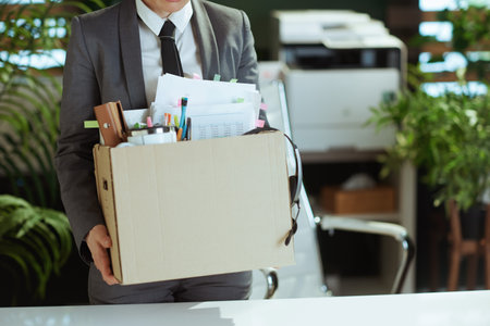 New job. Closeup on modern female employee in modern green office in gray business suit with personal belongings in cardboard box.の写真素材