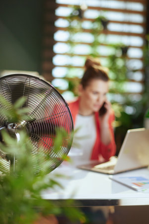 Sustainable workplace. Closeup on smiling modern accountant woman at work with electric fan and laptop.の写真素材