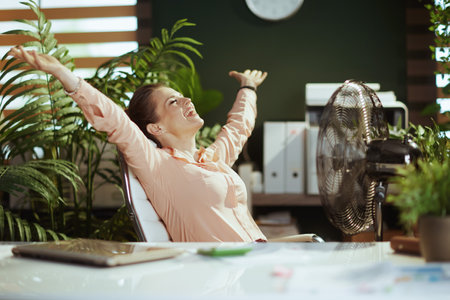 Sustainable workplace. happy modern bookkeeper woman in modern green office with electric fan and laptop.の写真素材