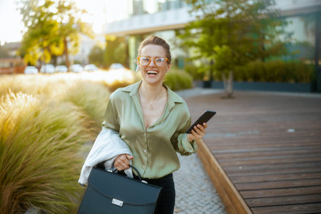 smiling modern middle aged woman employee in business district in green blouse and eyeglasses with smartphone and briefcase walking.の写真素材
