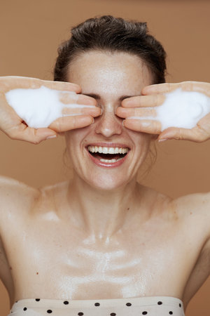happy middle aged woman with foaming facial cleanser washing face isolated on beige background.