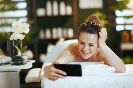 Healthcare time. smiling modern 40 years old woman in spa salon talking video call and laying on massage table.の写真素材