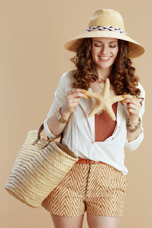 Beach vacation. smiling stylish middle aged woman in white blouse and shorts isolated on beige with sea star, straw bag and straw hat.の写真素材
