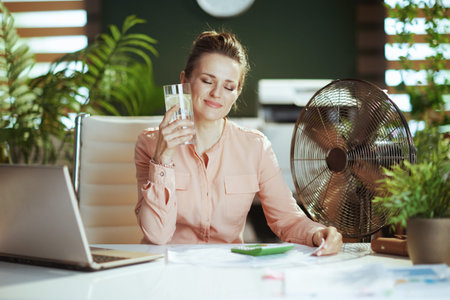 Sustainable workplace. happy modern woman worker at work with electric fan, laptop, cup of water and lemon.の写真素材