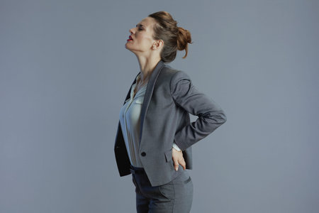 stressed stylish middle aged woman employee in gray suit having back pain isolated on gray...