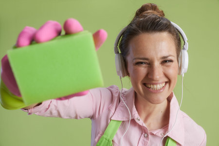 Spring cleaning. smiling modern woman in green apron and rubber gloves isolated on green background with headphones and cleaning sponge.の写真素材