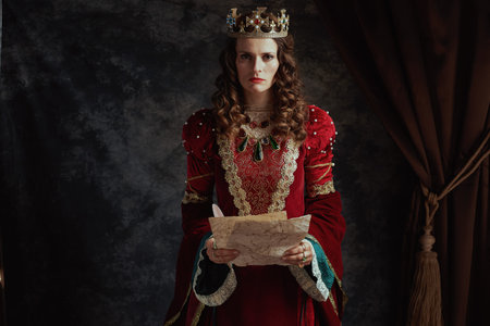 mad medieval queen in red dress with parchment and crown on dark gray background.の写真素材