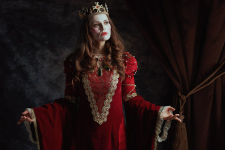 medieval queen in red dress with white makeup and crown on dark gray background.