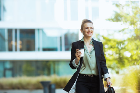 happy modern middle aged woman employee near office building in black jacket with cup of coffee walking.の写真素材